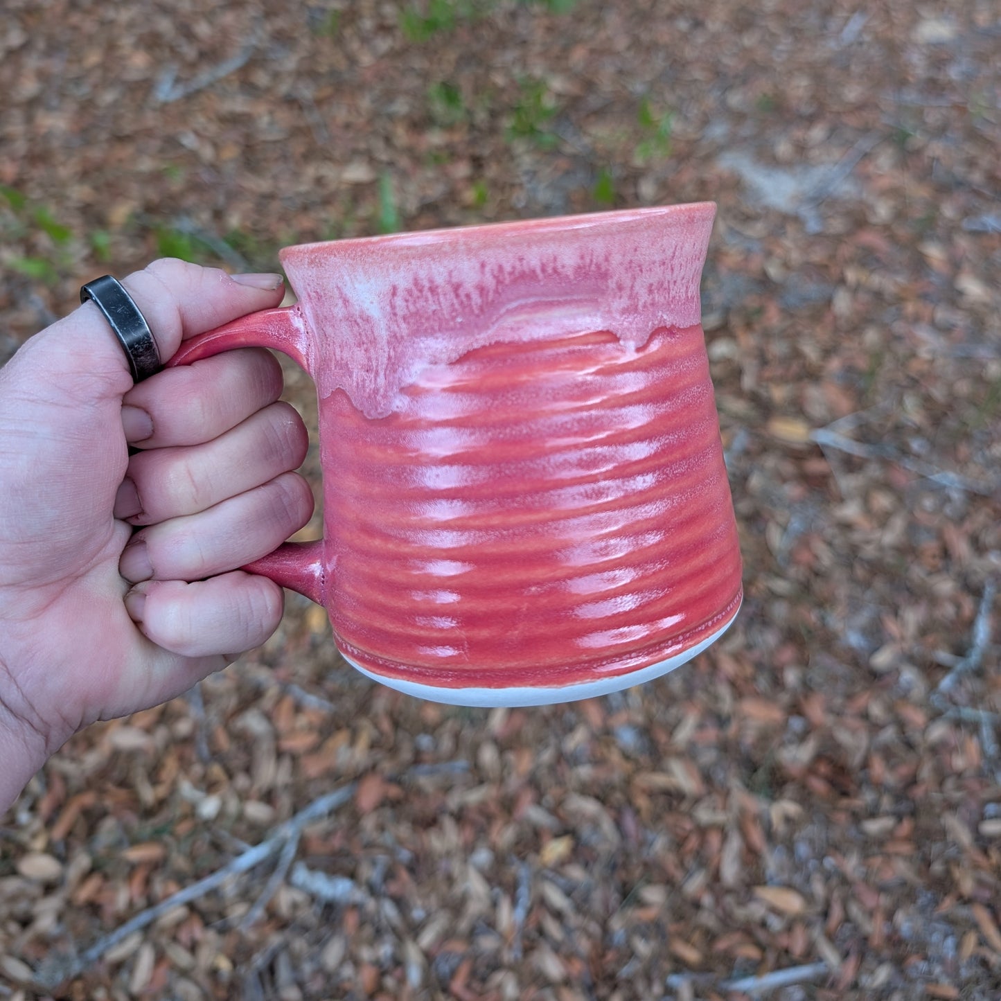 A hand holding a pinkish-red glazed ceramic mug outdoors against a blurred background of brown leaves and small green plants.