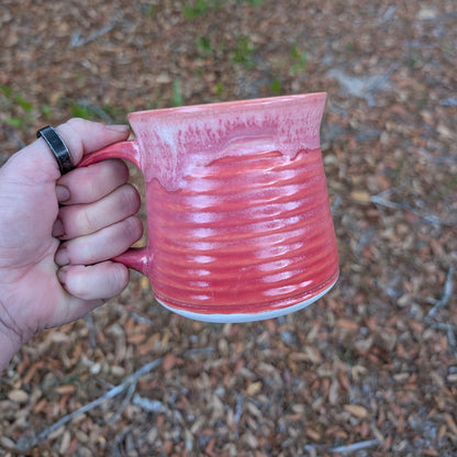 A hand holding a pinkish-red glazed ceramic mug outdoors against a blurred background of brown leaves and small green plants.