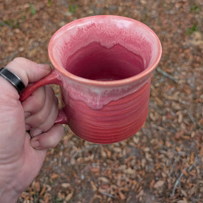 A hand holding a handmade ceramic mug with a pink and red glaze, set against a background of dry leaves and dirt.