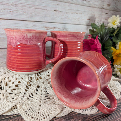 Three pinkish-red glazed ceramic mugs with ribbed handles, arranged on a white crocheted doily against a rustic white wood plank background with flowers in the corner.