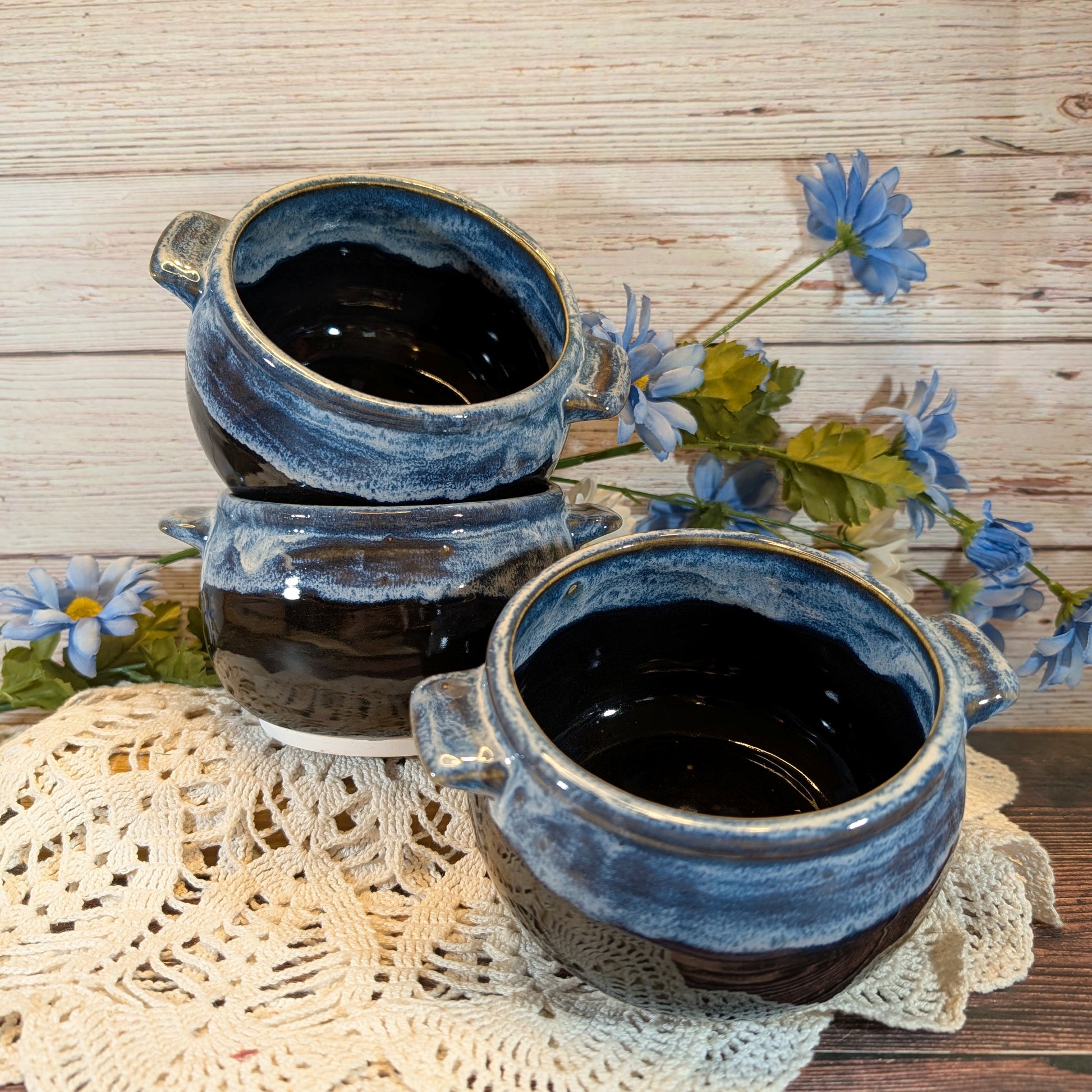 Three small ceramic bowls with a rustic blue and black glazed finish, arranged on a cream lace doily with blue flowers against a light wood plank background.