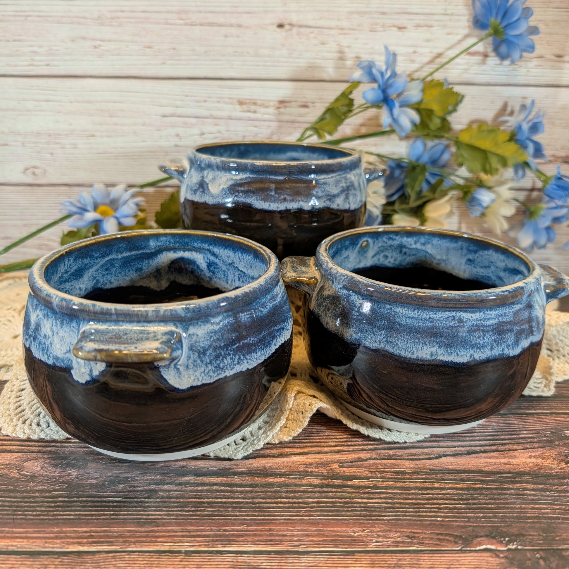 Three dark glazed ceramic bowls with blue-rimmed edges and small side handles, arranged on a wooden surface with blue flowers in the background.