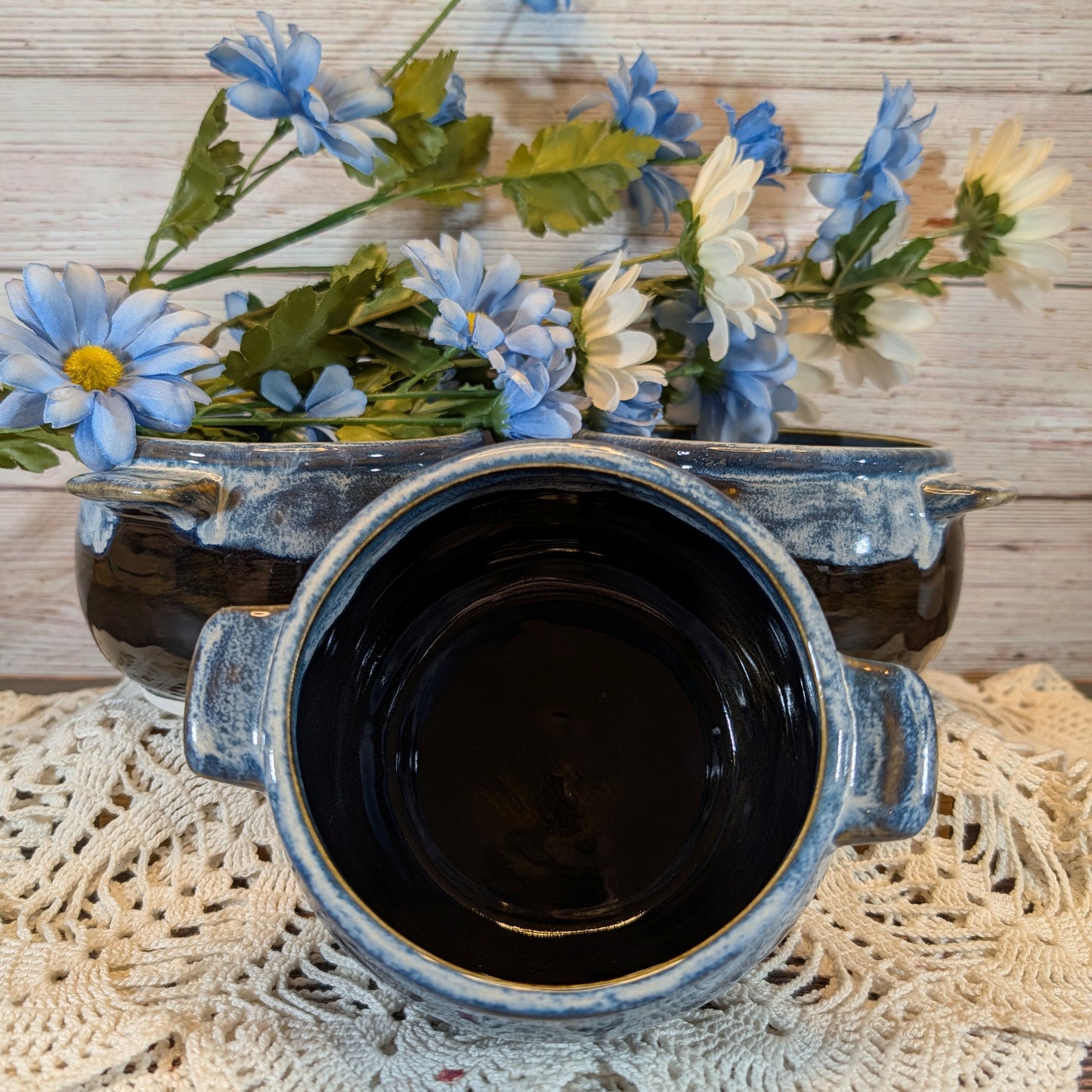 A rustic ceramic pot with a dark glazed interior and a weathered blue-gray exterior, styled on a lace doily with blue and white flowers in the background.