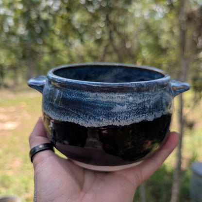 A handmade dark glazed ceramic pot with two side handles, held in a hand against a blurred outdoor background of trees and grass.