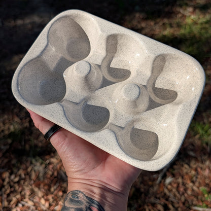 A hand holds a speckled light gray ceramic egg tray outdoors, with dry leaves and grass visible in the blurred background.