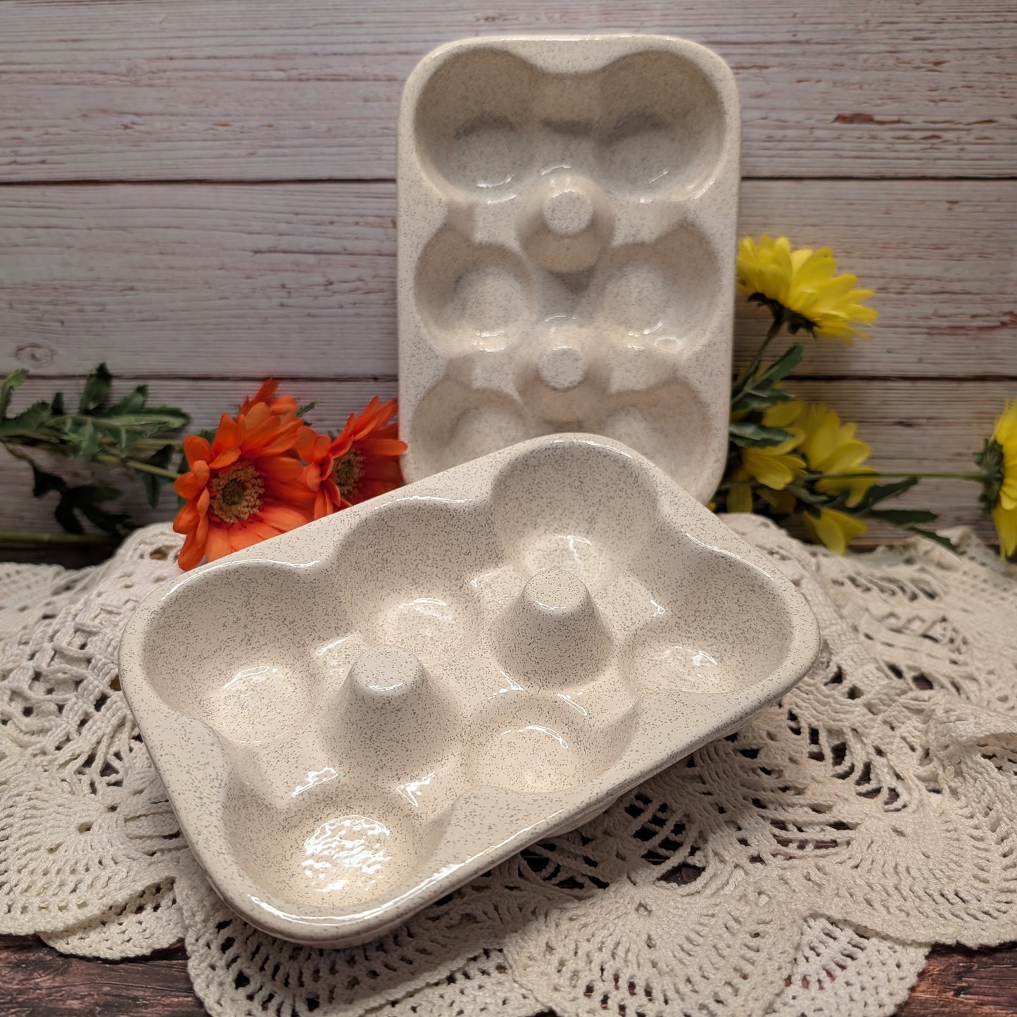 Two speckled ceramic egg trays,  displayed on a white lace doily with orange and yellow flowers against a rustic wood plank background.