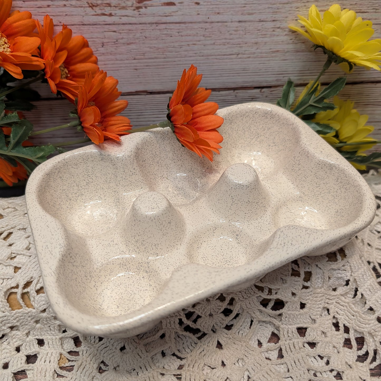 A speckled beige ceramic egg tray with six compartments, styled on a white lace doily with orange and yellow flowers against a rustic wooden background.