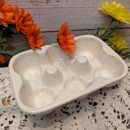 A speckled beige ceramic egg tray with six compartments, styled on a white lace doily with orange and yellow flowers against a rustic wooden background.