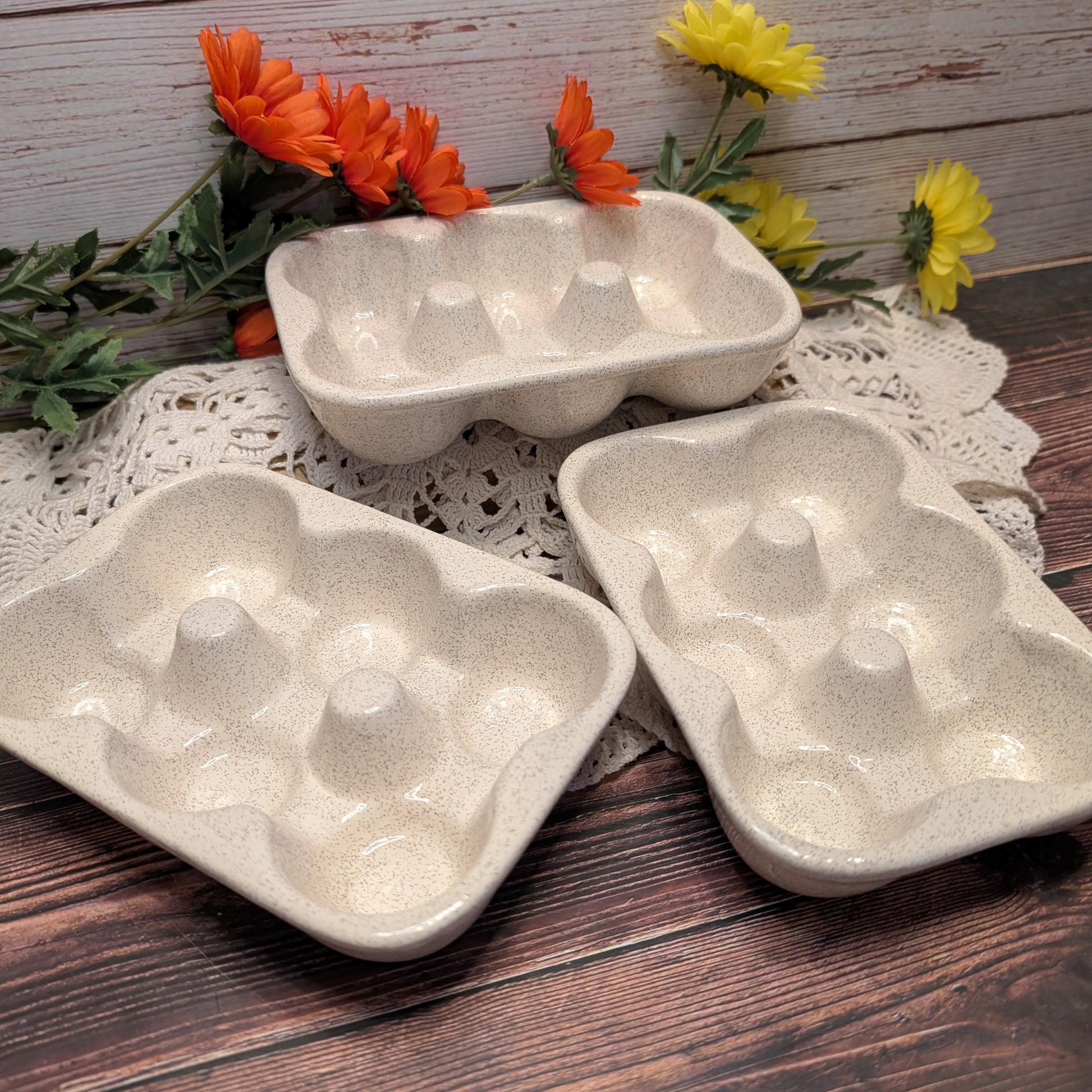 Three speckled ceramic egg trays arranged on a lace doily over a dark wood surface, with orange and yellow flowers in the background.