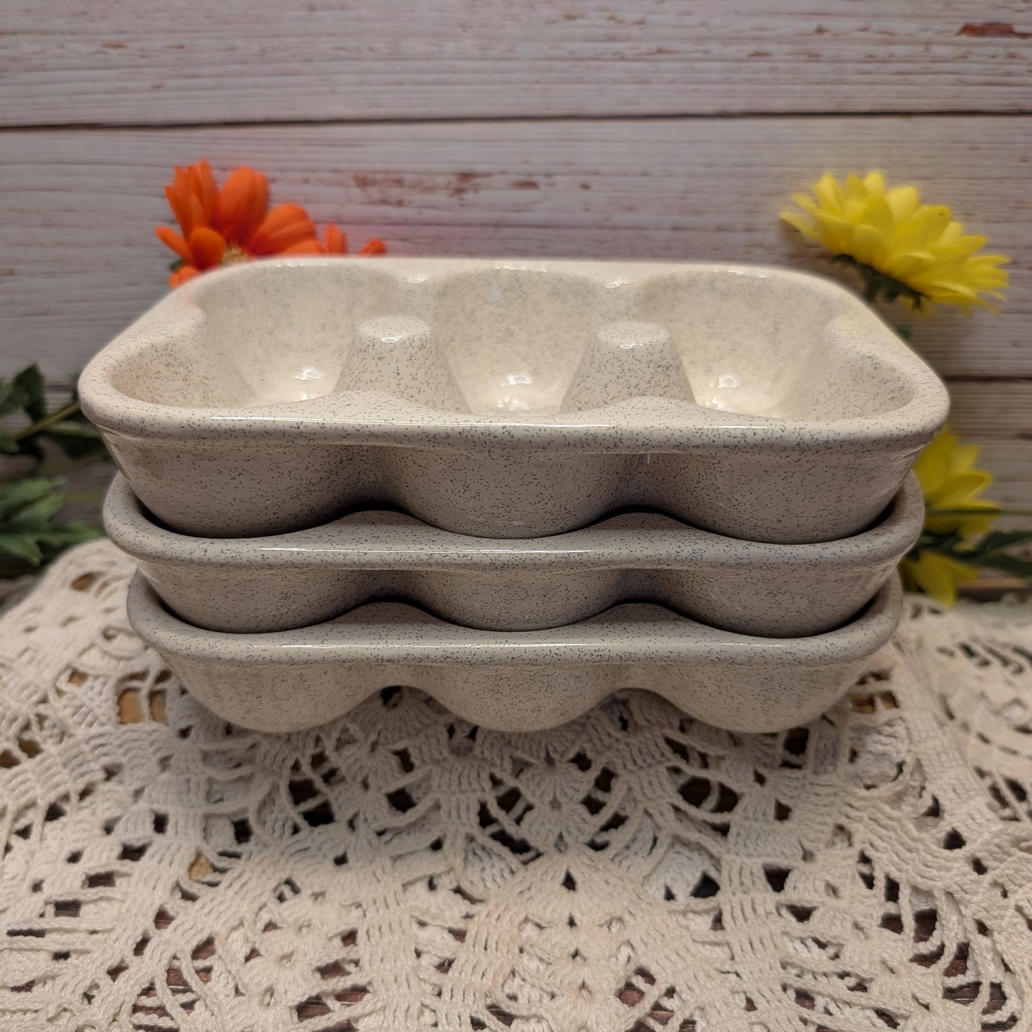 Three speckled beige ceramic egg trays stacked on a white lace doily, with orange and yellow flowers against a rustic whitewashed wood background.