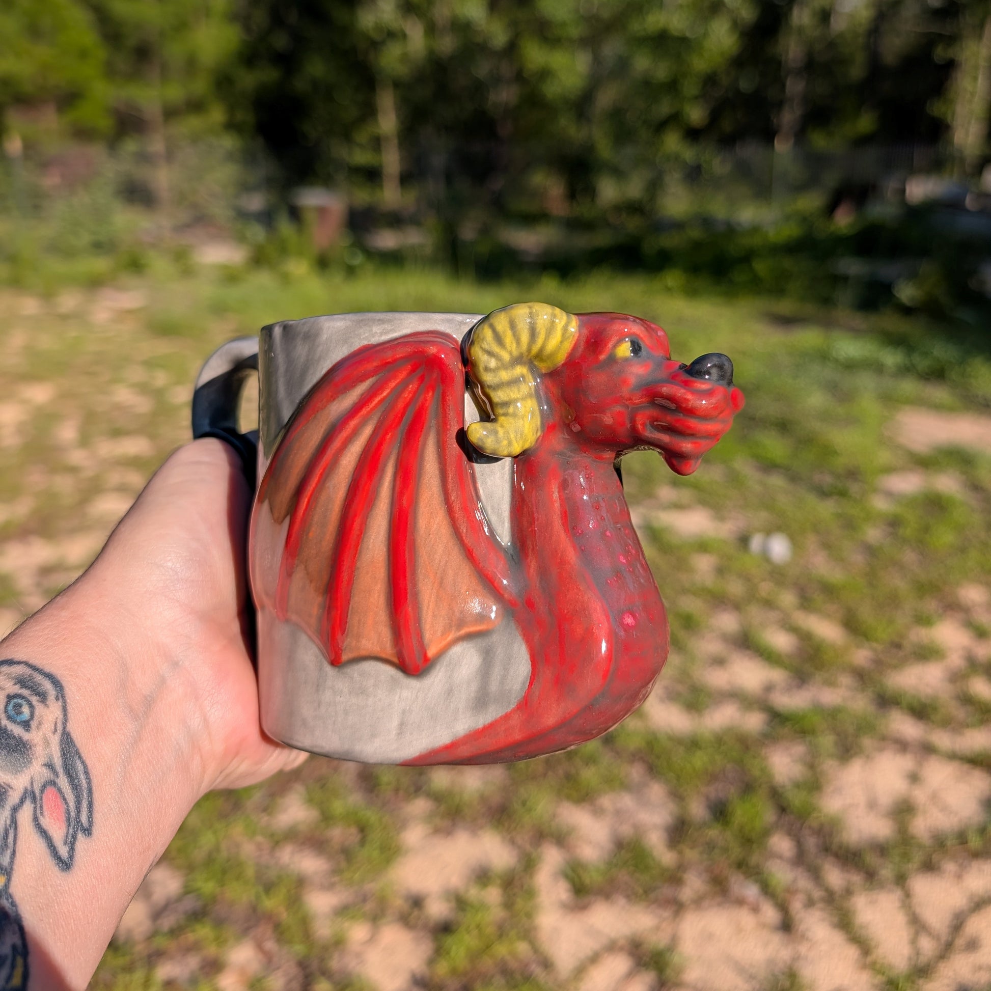 A hand holds a ceramic mug shaped like a red dragon with yellow horns and wings, set against a blurred outdoor background of grass and trees.