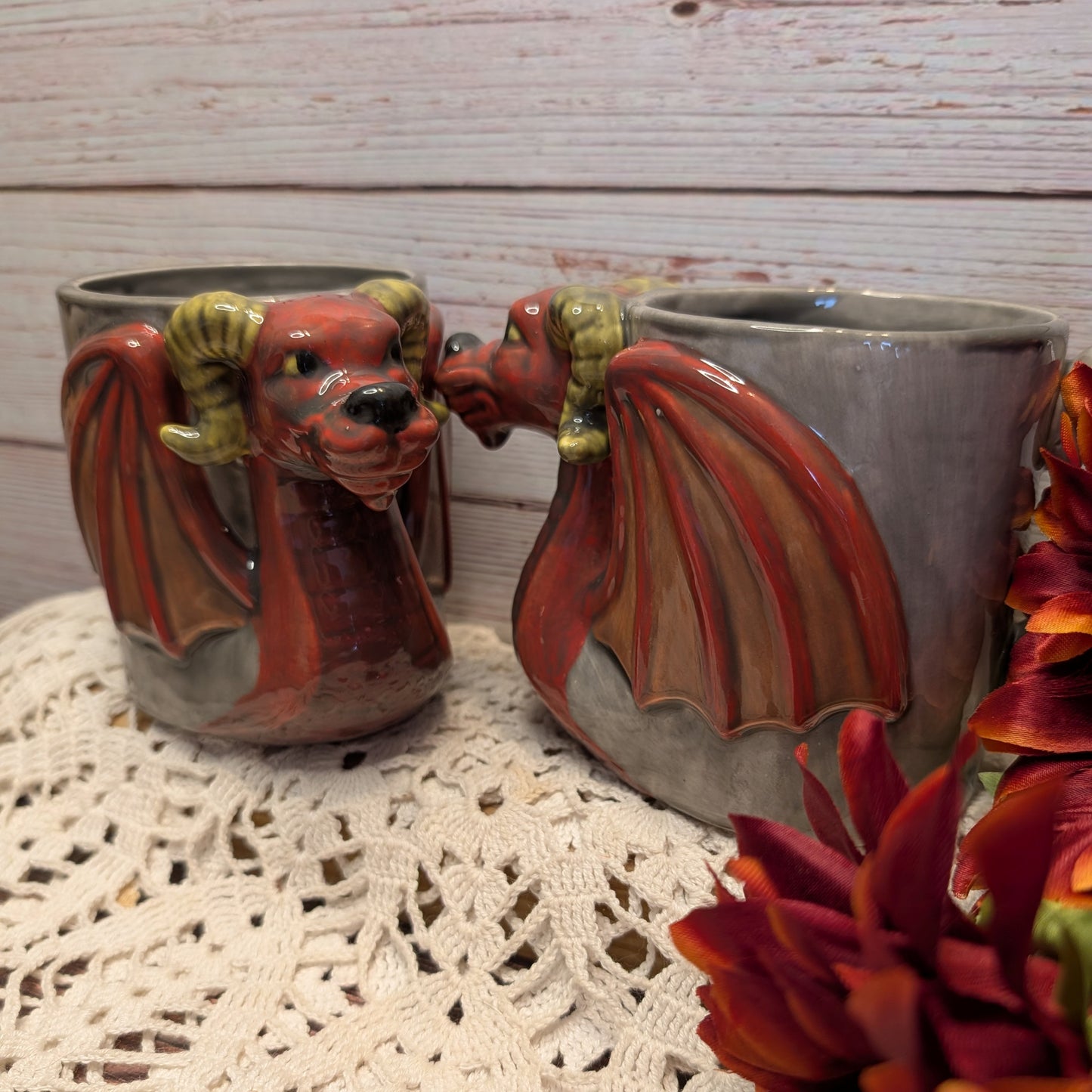 Two ceramic dragon mug shaped like red and brown dragons with green horns, displayed on a white lace doily against a whitewashed wood background.