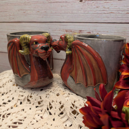 Two ceramic dragon mug shaped like red and brown dragons with green horns, displayed on a white lace doily against a whitewashed wood background.
