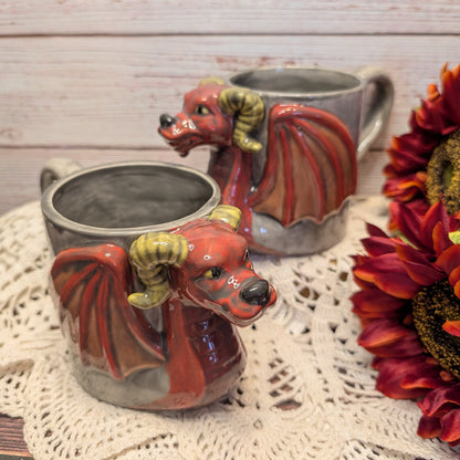 Two ceramic dragon mugs with red bodies, gray wings, and yellow horns, styled on a white lace doily with sunflowers against a light wood background.