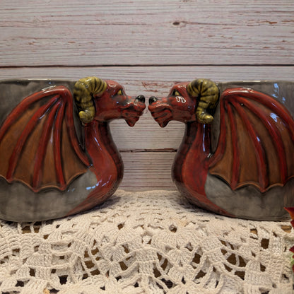 A pair of decorative ceramic dragon mugs shaped like facing dragons with red wings, brown bodies, and green horns, displayed on a white crocheted doily against a light wood plank background.