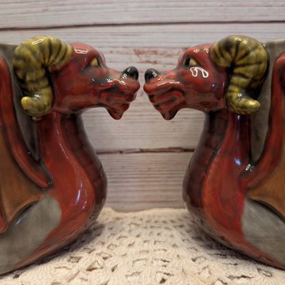 A pair of glazed ceramic dragon mugs facing each other, featuring reddish-brown bodies, olive-green horns, and gray backs, displayed on a textured cream surface against a light wood plank background.