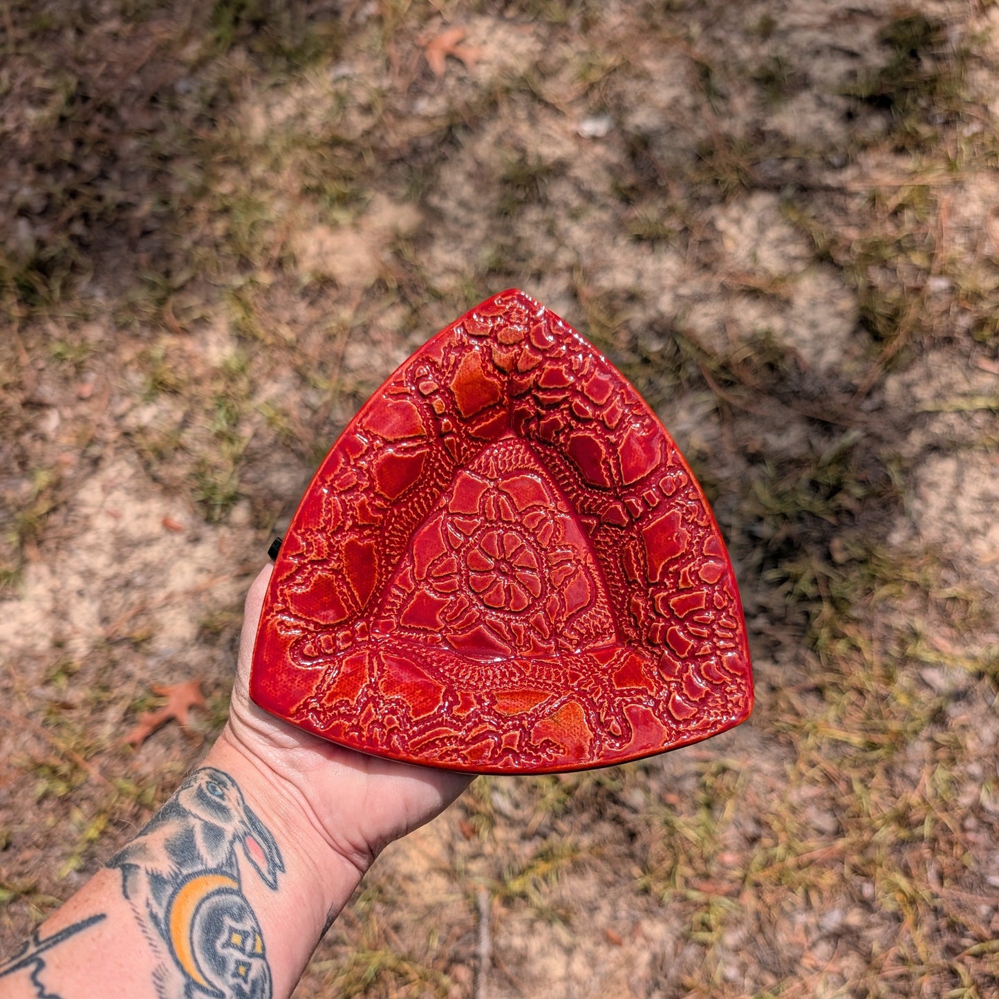 A hand holding a red textured ceramic triangular dish outdoors on a sandy ground.
