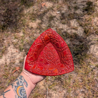 A hand holding a red textured ceramic triangular dish outdoors on a sandy ground.