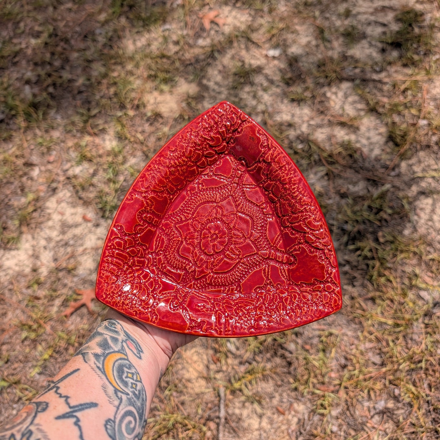 A red triangular ceramic plate with a raised floral mandala pattern, held by a tattooed arm against a blurry outdoor ground background.