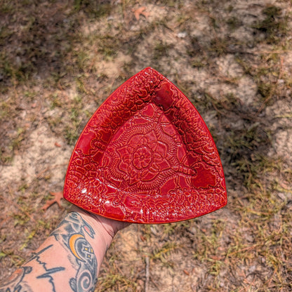 A red triangular ceramic plate with a raised floral mandala pattern, held by a tattooed arm against a blurry outdoor ground background.