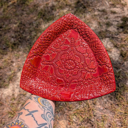 A red textured ceramic plate with a floral embossed design, held by a tattooed arm against a blurry outdoor ground background.