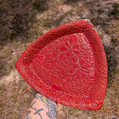 A red textured ceramic plate with a raised floral pattern, held by a tattooed arm against an outdoor background of dirt and grass.