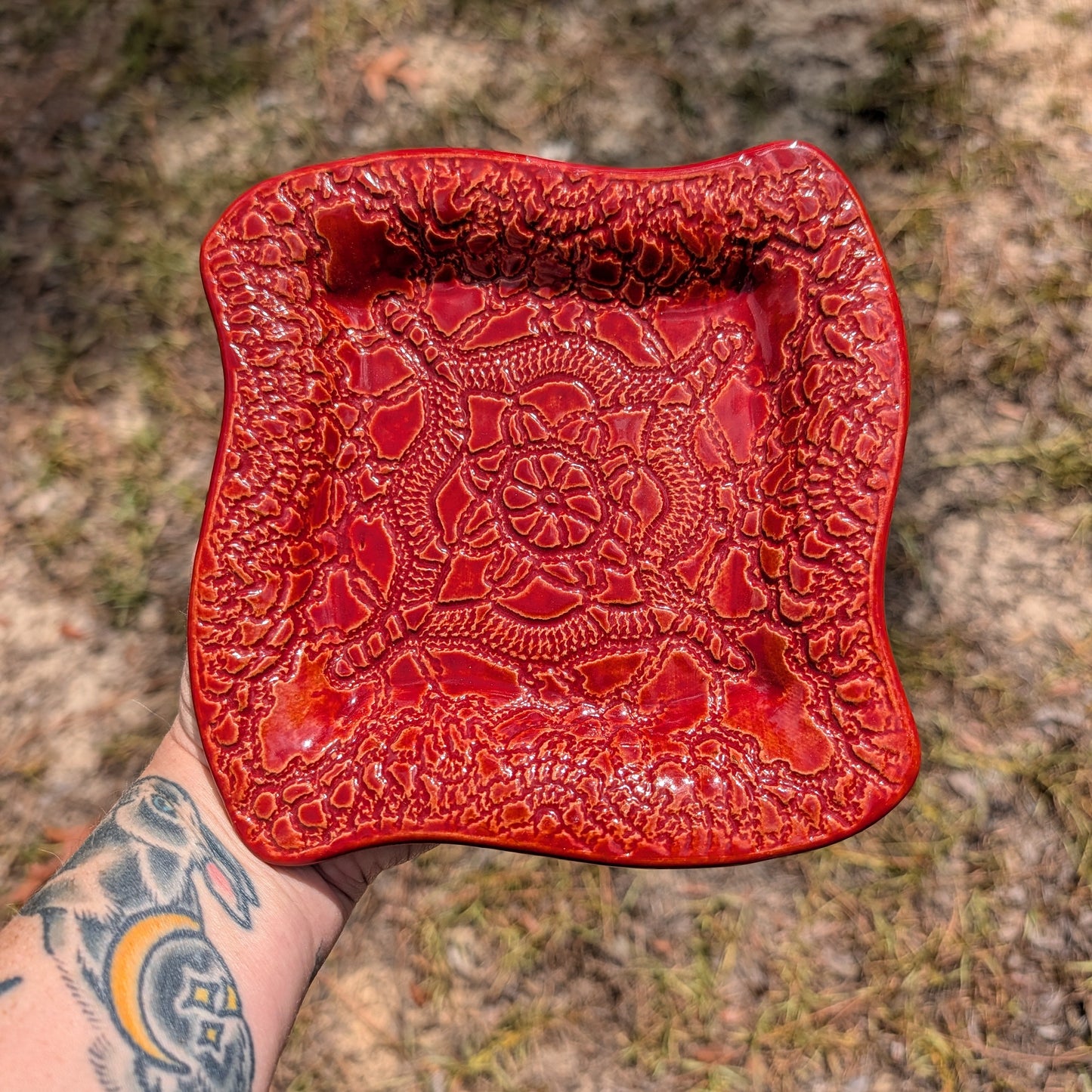 A hand with a colorful tattoo holds a square, wavy-edged red ceramic dish with an embossed floral mandala pattern, set against an outdoor grassy background.