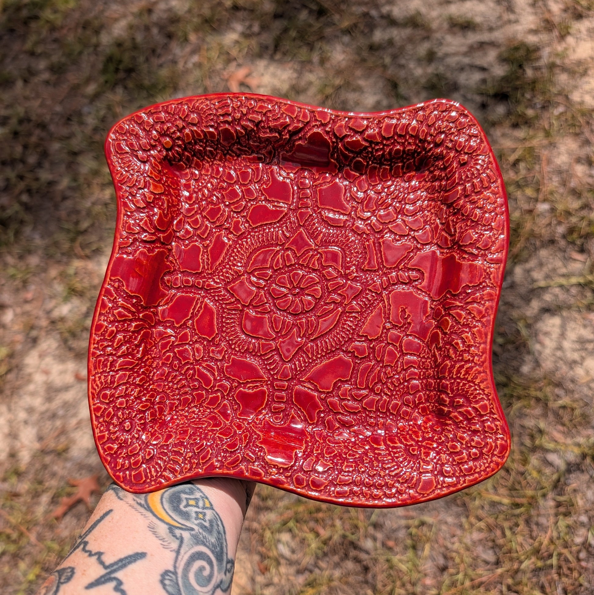 A square red ceramic plate with a raised floral pattern, held by a tattooed arm against an outdoor background of dirt and grass.