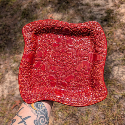 A square red ceramic plate with a raised floral pattern, held by a tattooed arm against an outdoor background of dirt and grass.