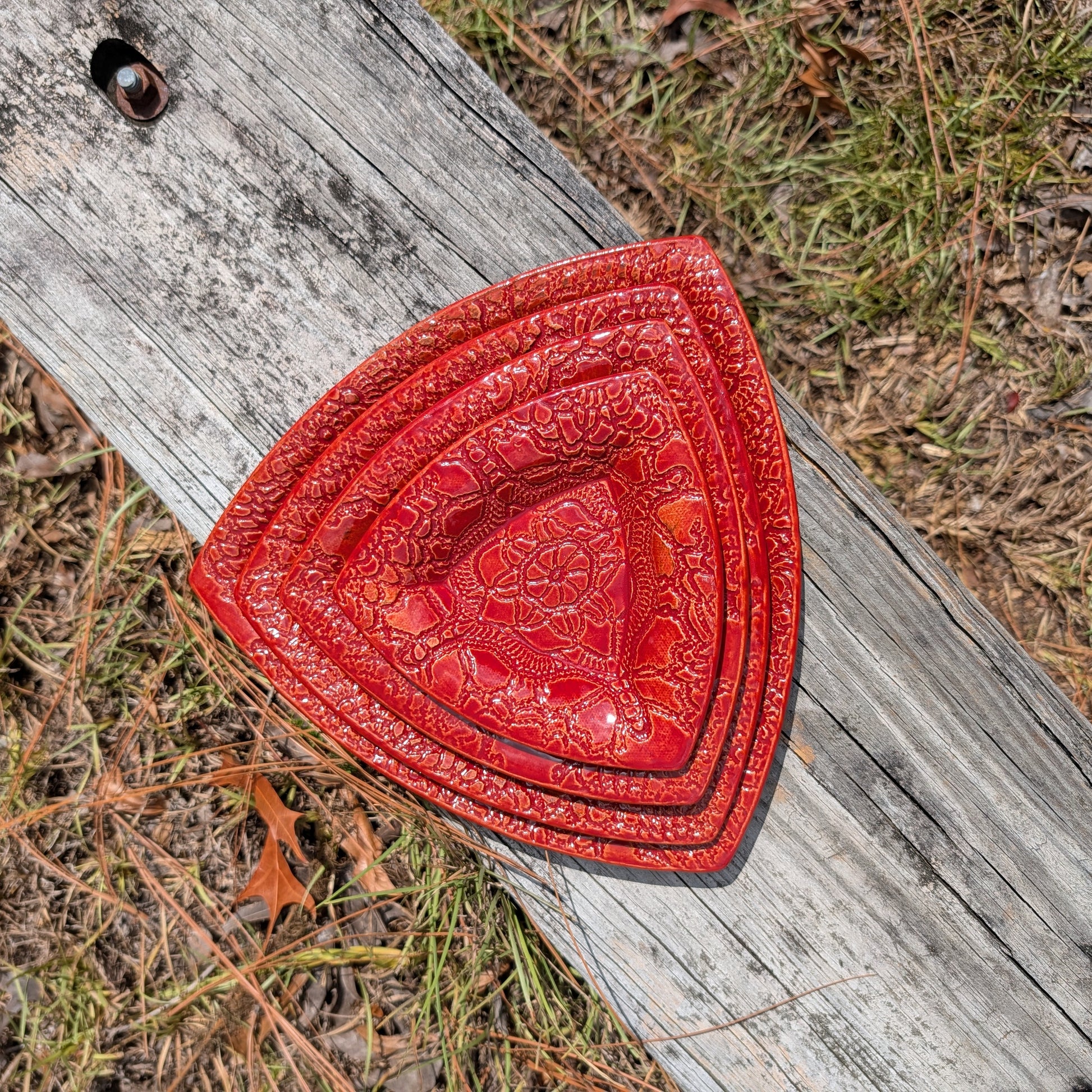 Four nested red ceramic trays with embossed floral patterns, displayed outdoors on a weathered wooden plank with grass and dry leaves in the background.