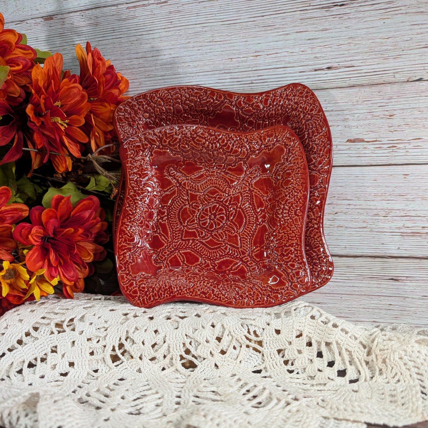 A square red ceramic plate with wavy edges and an embossed floral pattern, styled on a white crocheted doily next to orange and red flowers against a whitewashed wood background.