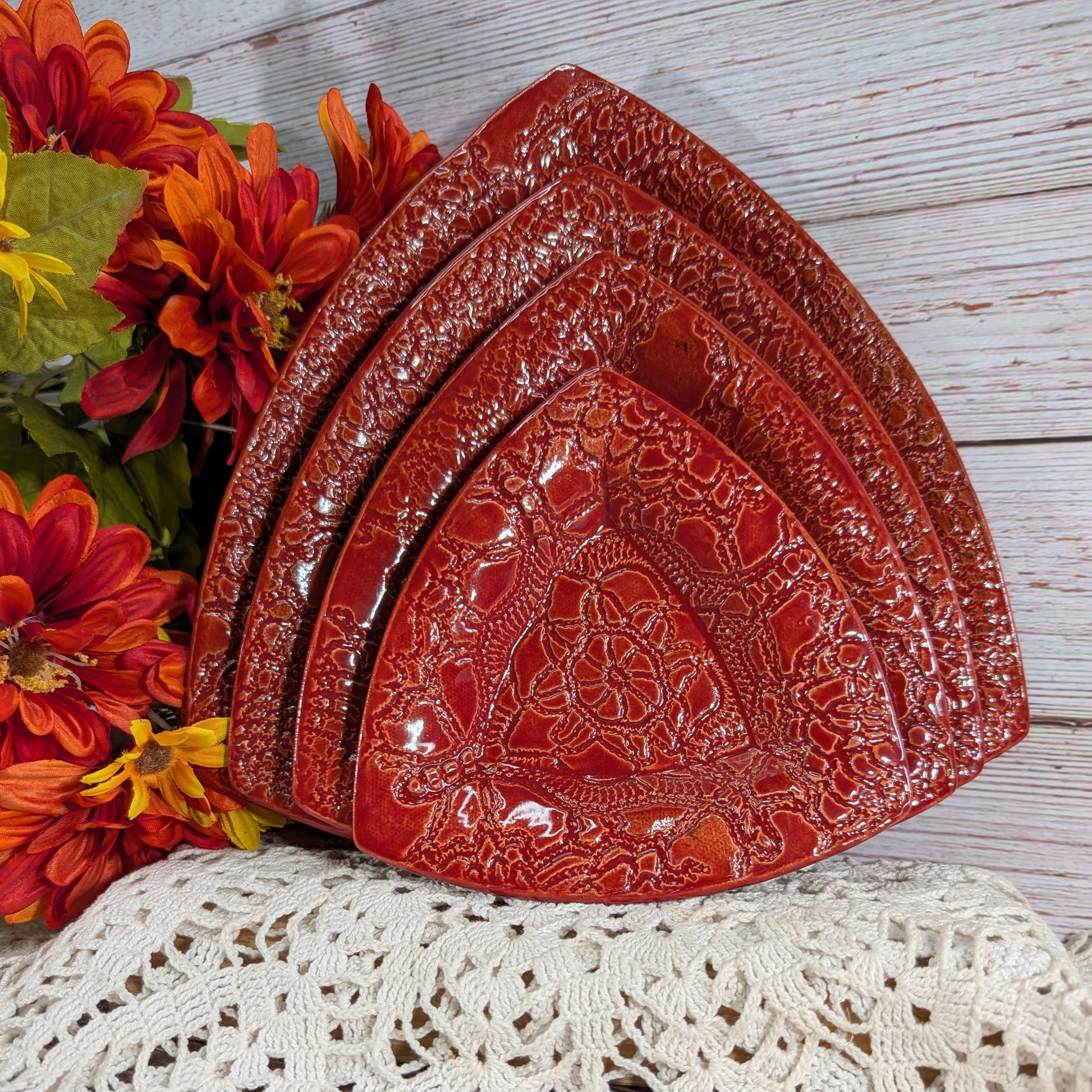 Four nested red ceramic triangular plates with embossed floral patterns, arranged on a white lace doily next to orange and red artificial flowers on a white wood background.
