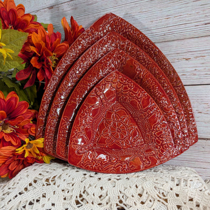 Four nested red ceramic triangular plates with embossed floral patterns, arranged on a white lace doily next to orange and red artificial flowers on a white wood background.