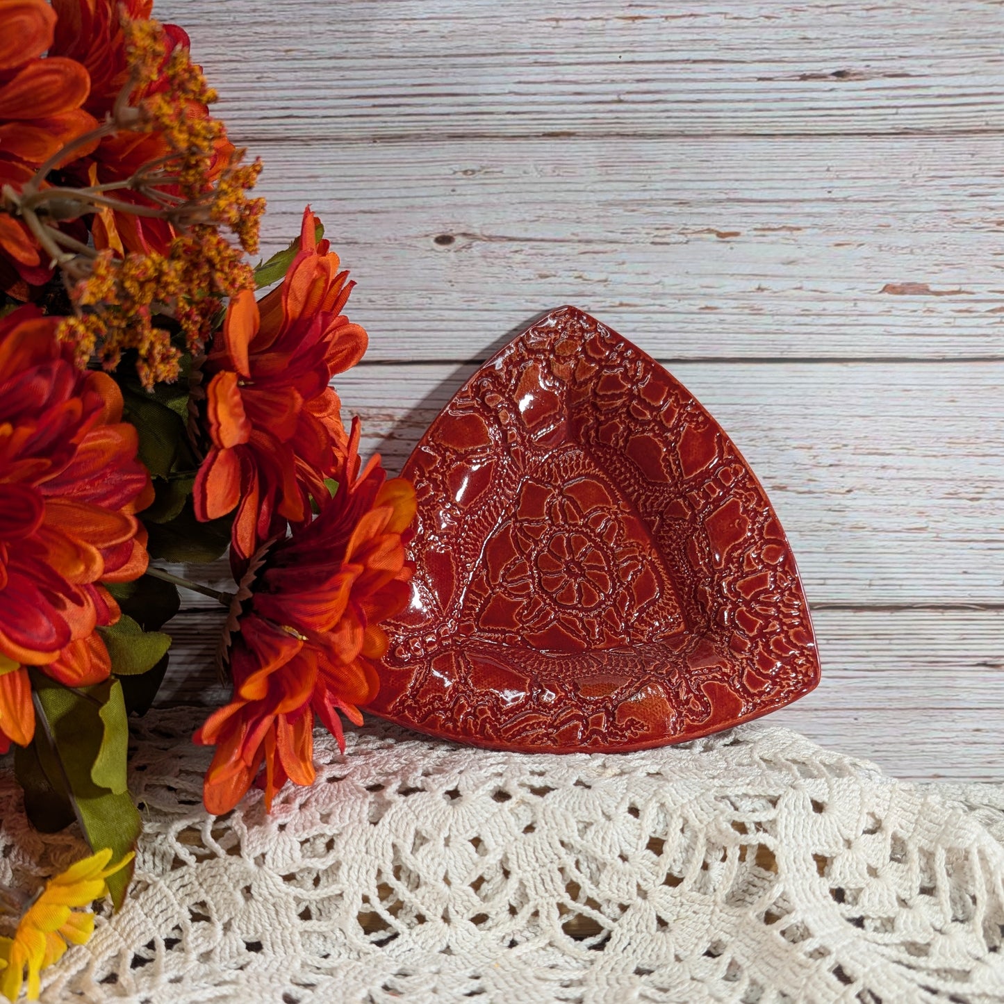 A small triangular red ceramic plate with embossed floral patterns, styled on a white lace doily next to orange flowers on a rustic white wood background.