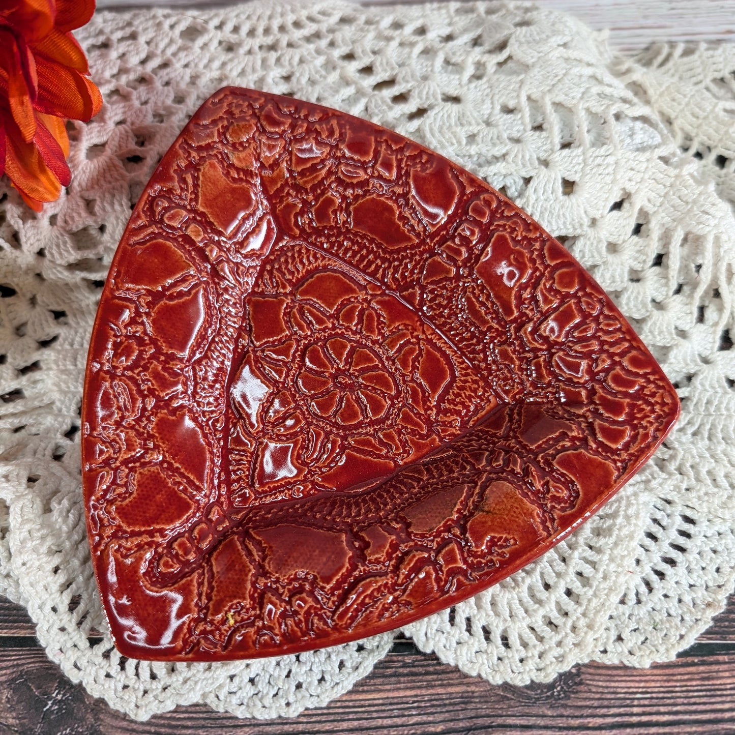 A glossy red ceramic dish with embossed floral and scrollwork patterns, styled on a white crocheted doily with a small orange flower in the corner.
