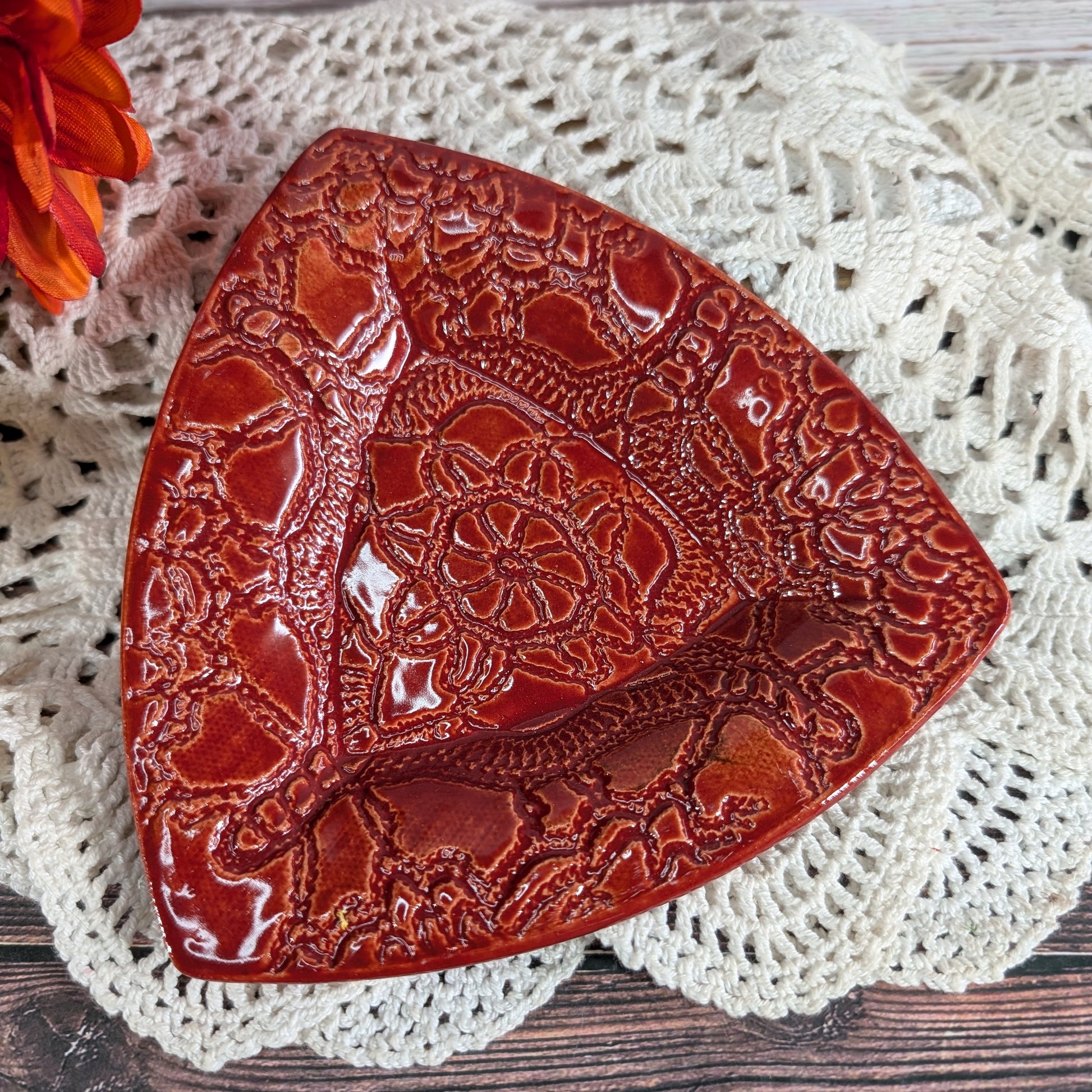 A glossy red ceramic dish with embossed floral and scrollwork patterns, styled on a white crocheted doily with a small orange flower in the corner.
