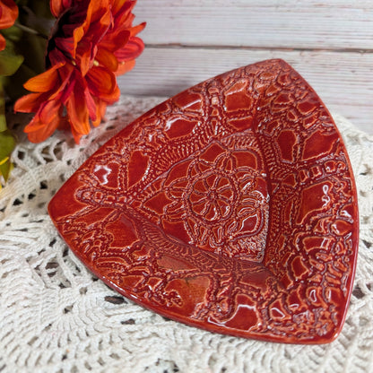 A glossy red ceramic dish with embossed floral and geometric patterns, styled on a white crocheted doily with orange flowers in the background.