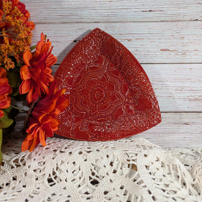 A red triangular ceramic plate with an embossed floral mandala pattern, styled on a white lace doily with orange flowers against a white wood plank background.