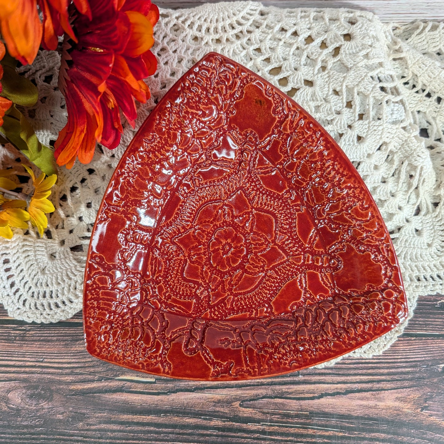 A glossy red ceramic serving platter with embossed floral and geometric patterns, styled on a white crocheted doily over a rustic wooden surface with orange flowers in the corner.