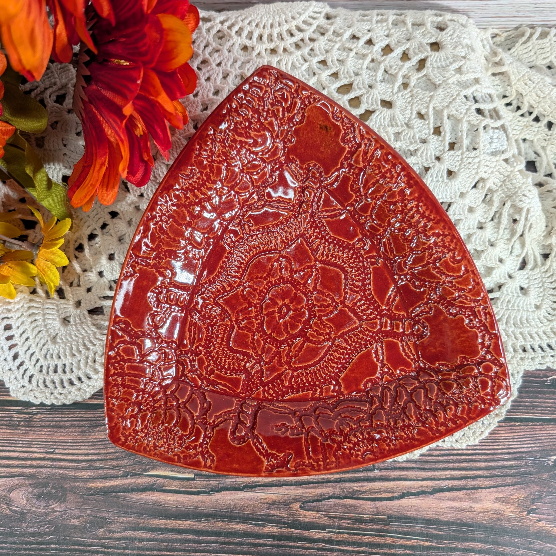 A glossy red ceramic serving platter with embossed floral and geometric patterns, styled on a white crocheted doily over a rustic wooden surface with orange flowers in the corner.
