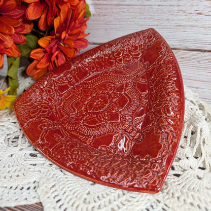 A glossy red ceramic dish with embossed floral and scroll patterns, styled on a white lace doily with orange flowers in the background.