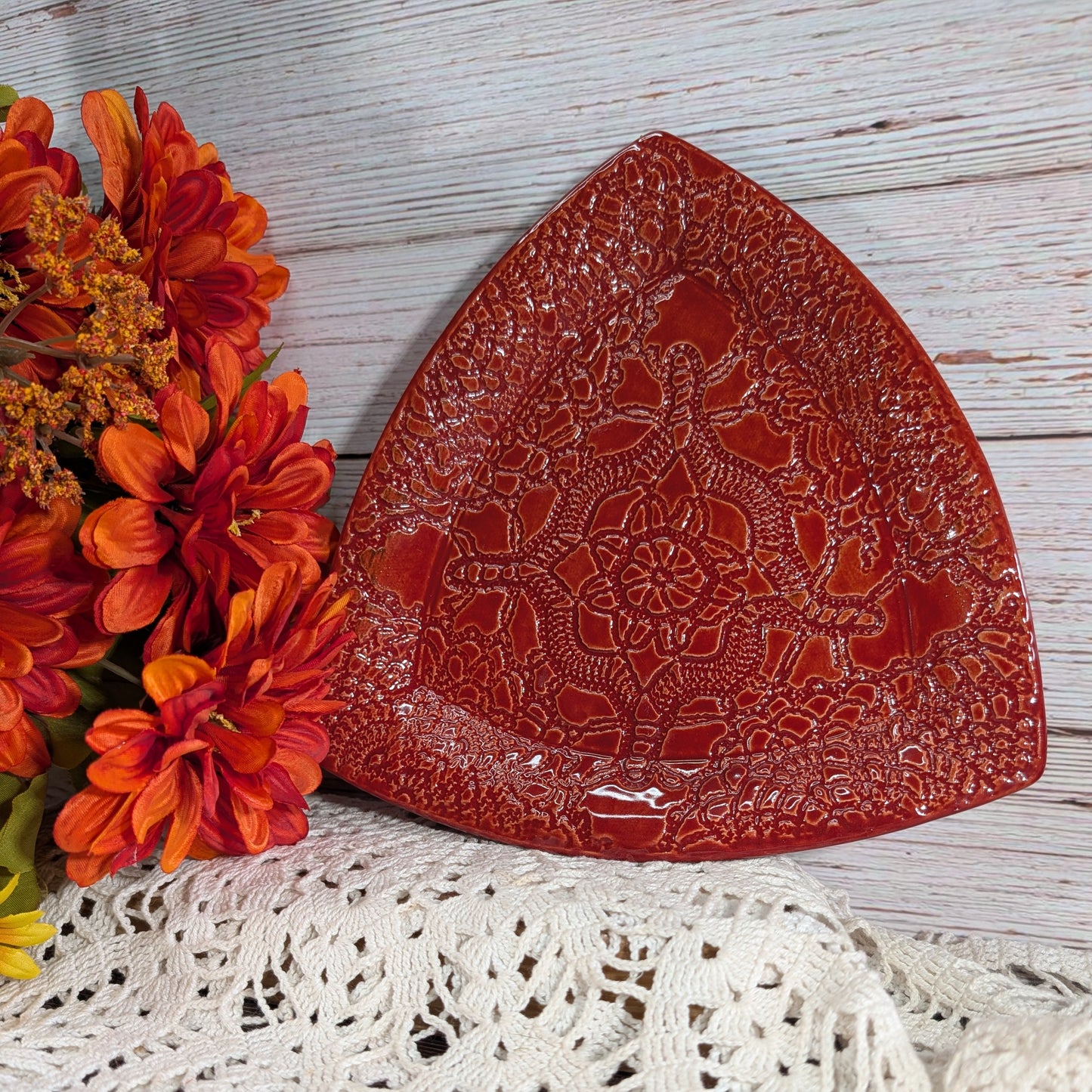 A red ceramic triangular serving dish with an embossed floral pattern, styled on a white lace doily next to orange and red flowers against a whitewashed wood background.