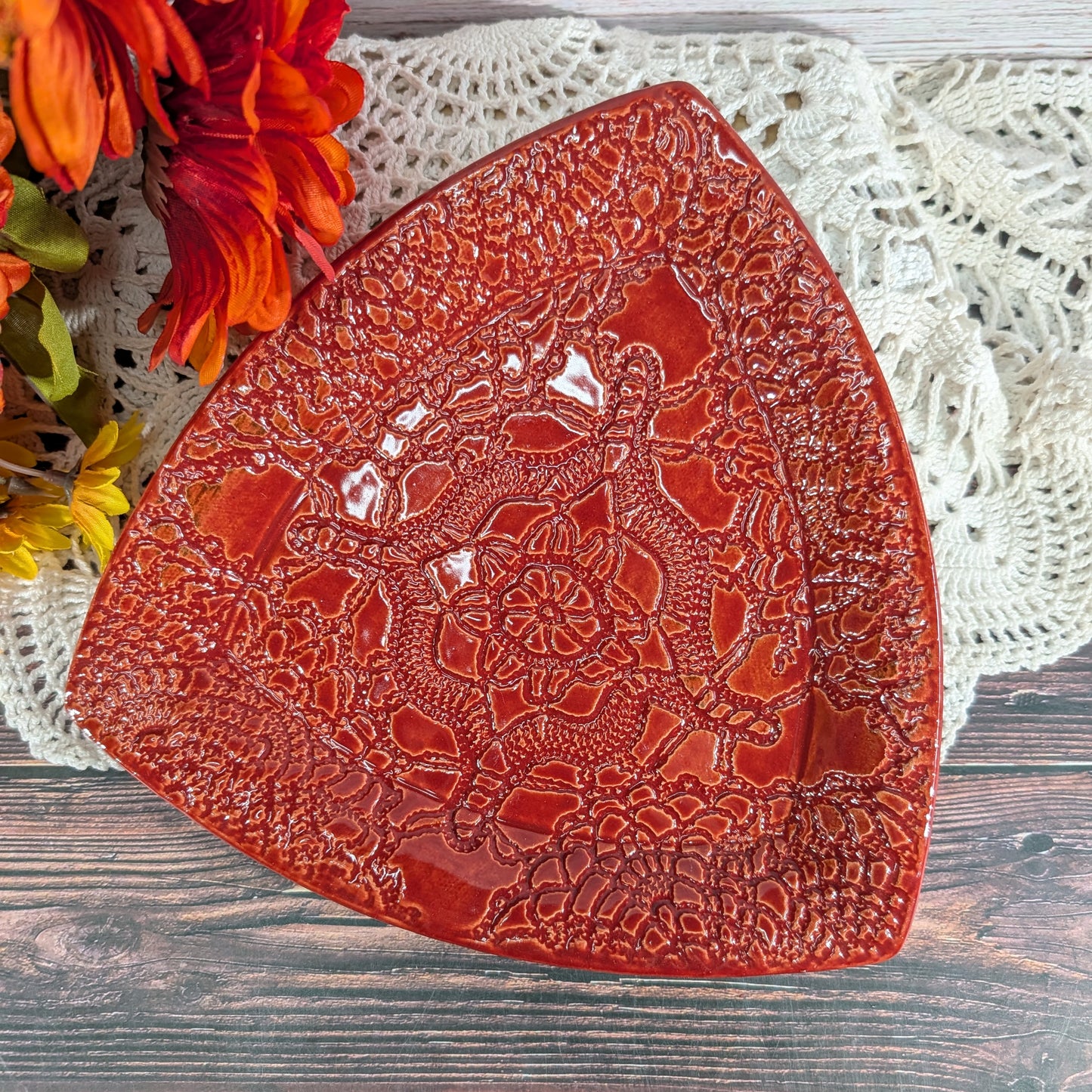 A red ceramic plate with an embossed floral lace pattern, displayed on a white crocheted doily over a dark wood surface with orange flowers in the corner.