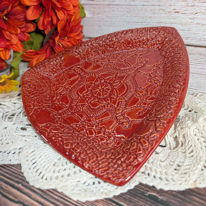 A red ceramic decorative plate with an embossed floral and geometric pattern, styled on a white crocheted doily with orange flowers in the background.