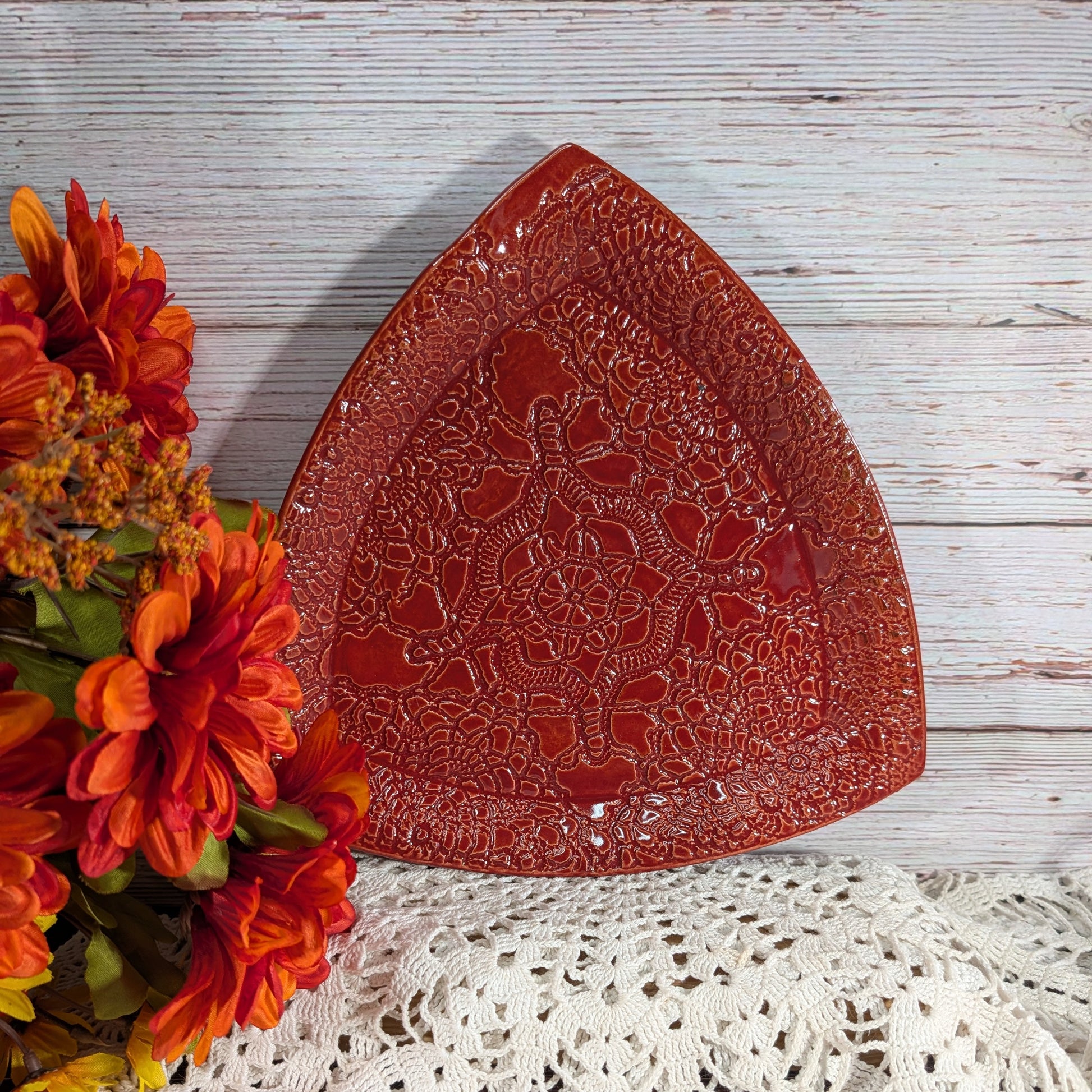 A red triangular ceramic plate with an embossed floral pattern, styled on a white wooden surface with a white lace doily and orange flowers.