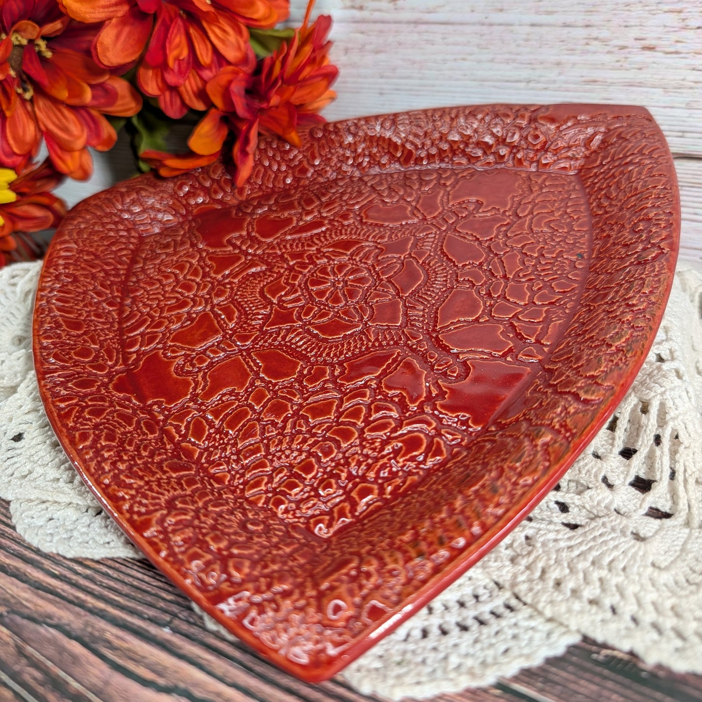 A triangular red ceramic plate with a raised floral lace-like pattern, styled on a white crocheted doily with orange flowers in the background.