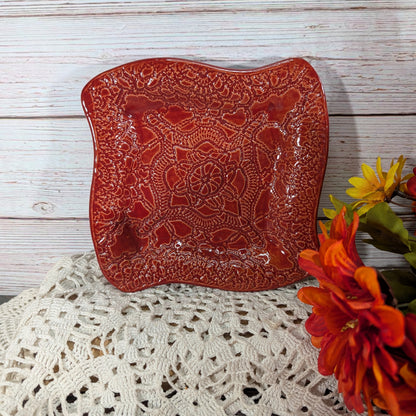 A square red ceramic plate with a raised floral mandala pattern, styled on a white lace doily against a whitewashed wood background with orange flowers on the side.