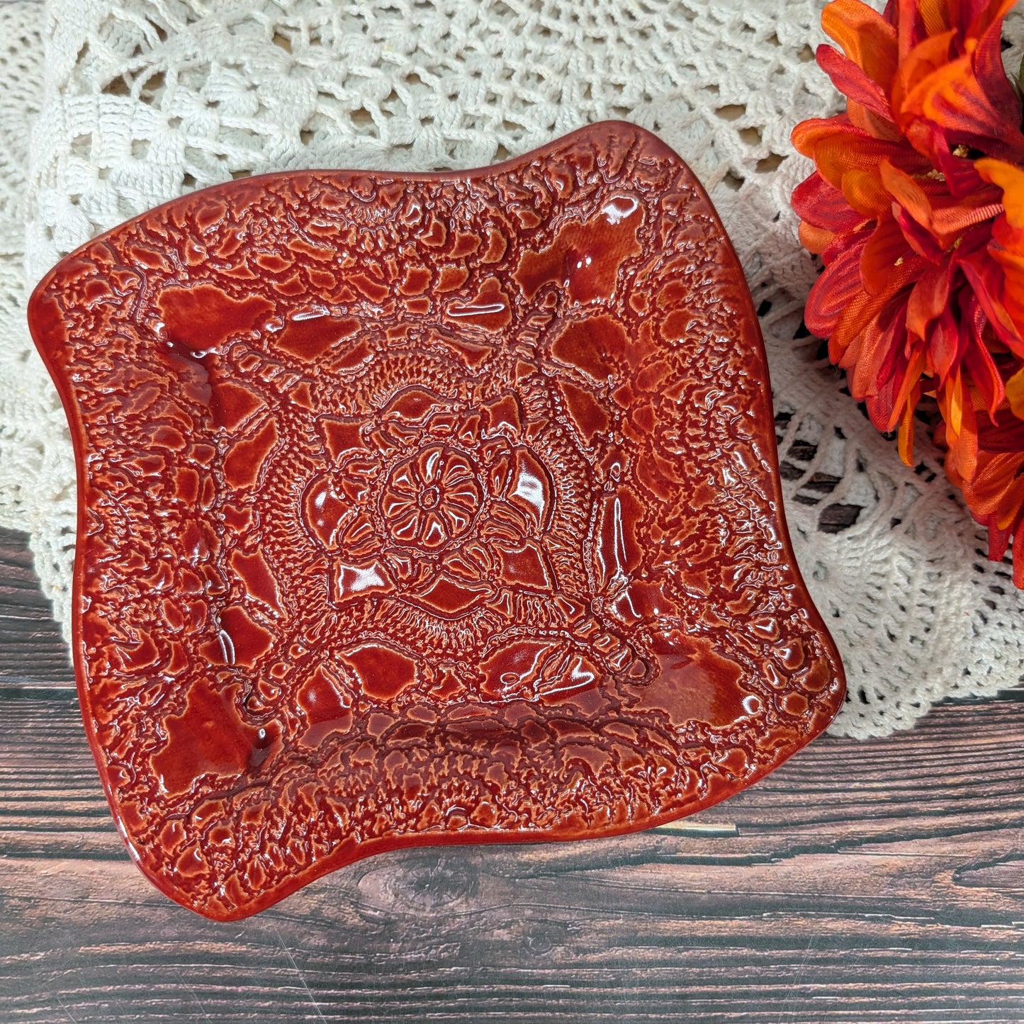 A square glossy red glass plate with a raised floral and geometric pattern, styled on a white lace doily over a dark wood surface next to orange flowers.