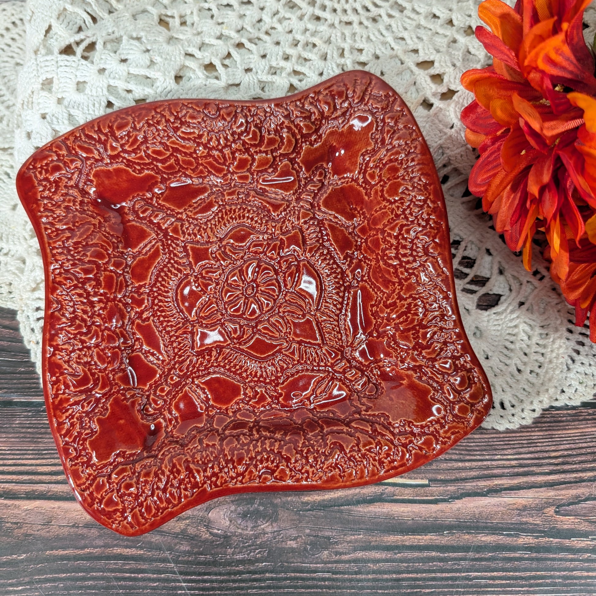 A square glossy red glass plate with a raised floral and geometric pattern, styled on a white lace doily over a dark wood surface next to orange flowers.