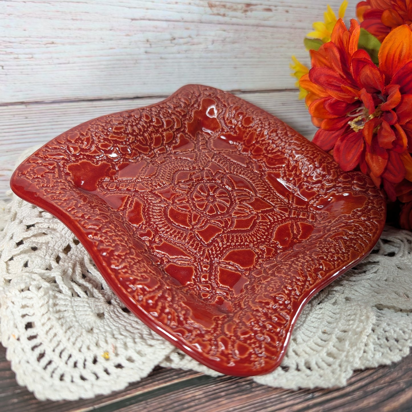 A glossy red square ceramic plate with embossed floral and scrollwork patterns, styled on a white crocheted doily with orange and yellow flowers in the background.
